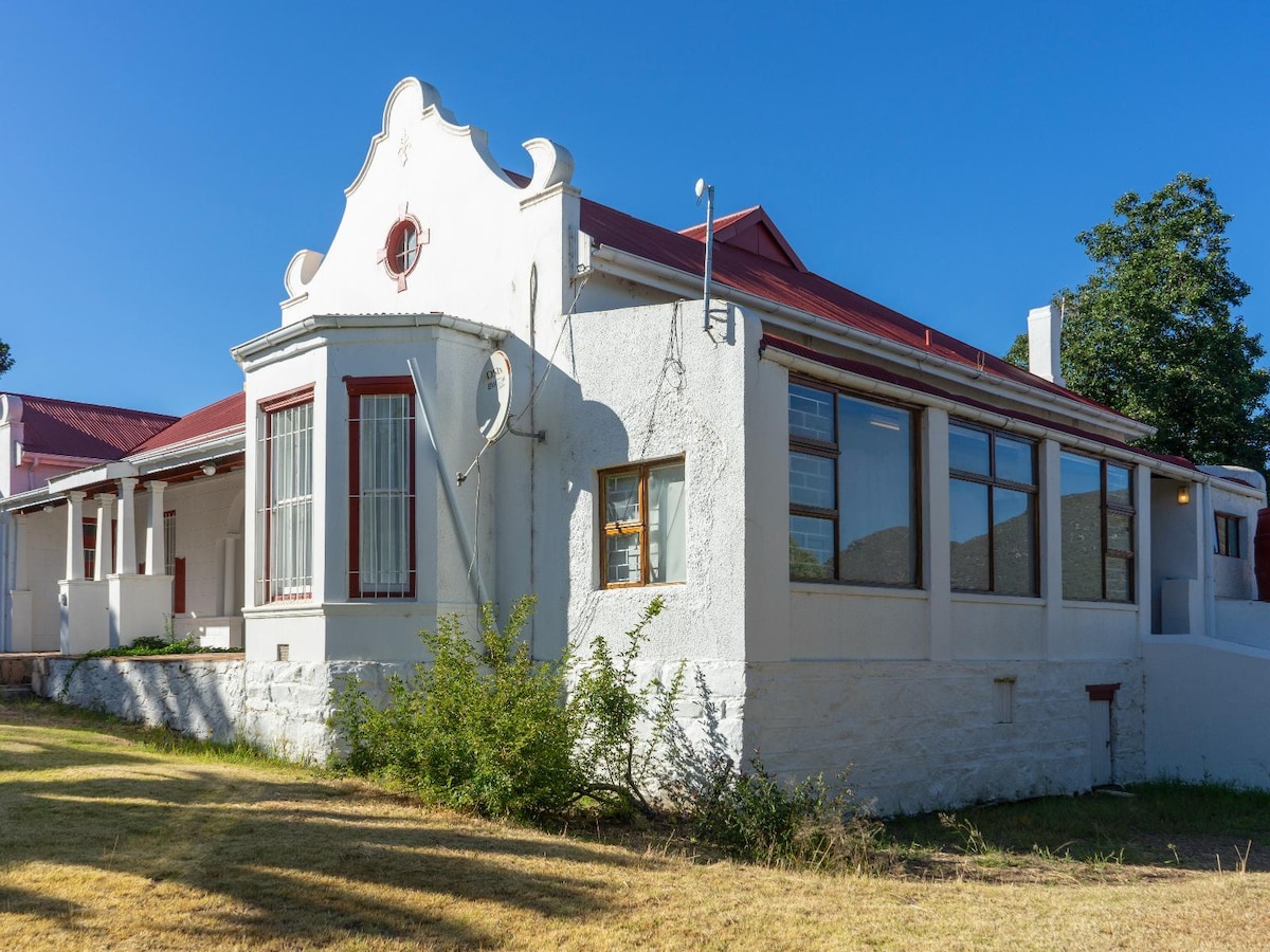 The exterior of the farmhouse is depicted, showcasing a white façade adorned with red windows and a distinctive gabled roof. A spacious garden area surrounds the building, with large windows allowing natural light into the interior.