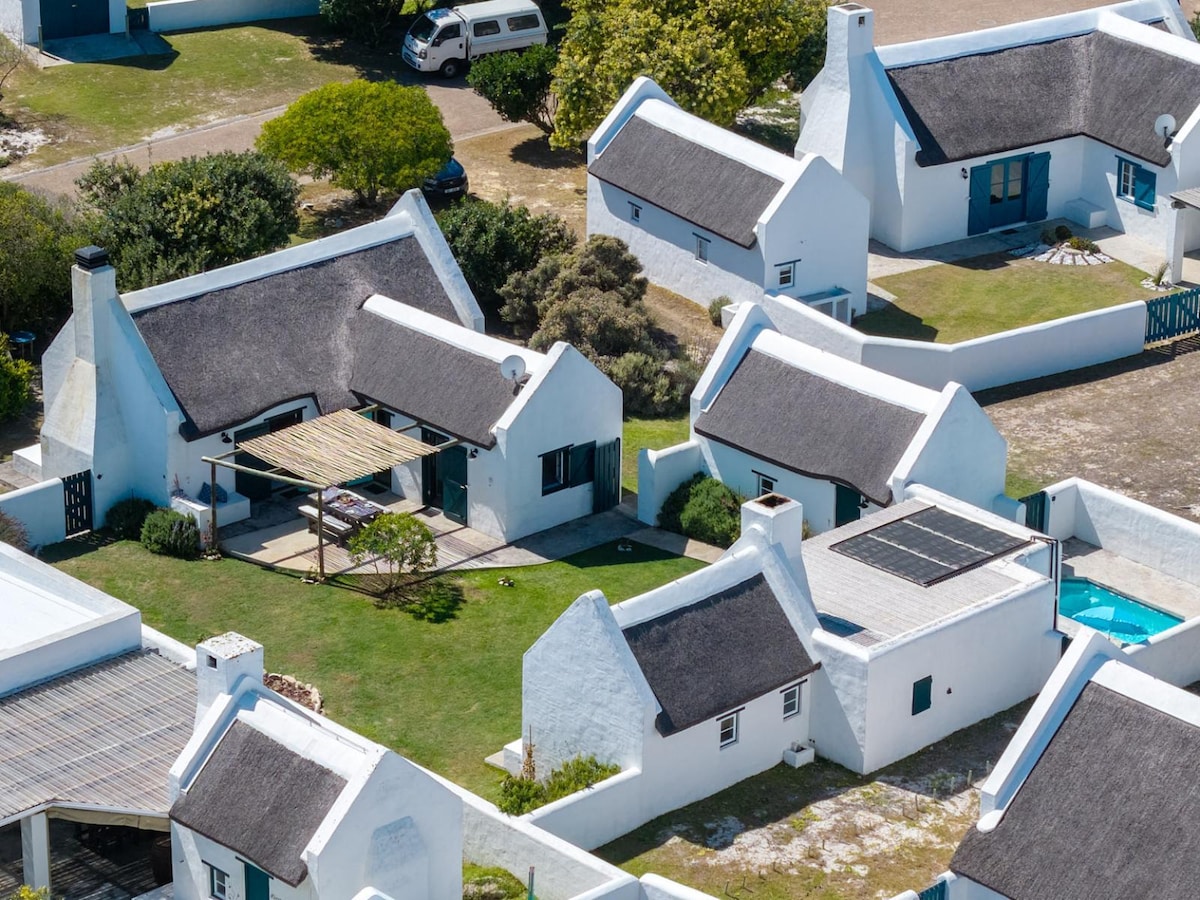 An aerial view captures the layout of Southern Hideaway Cottage and surrounding buildings, each featuring thatched roofs. Green lawns and landscaped shrubs complement the white structures, while a solar panel is visible on one roof. A small pool adds a serene touch to the outdoor space.