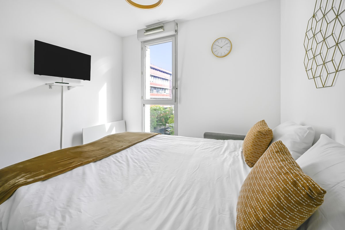 A bright bedroom features a spacious bed adorned with decorative pillows. A wall-mounted television is positioned opposite the bed. Natural light enters through a large window, complemented by a minimalist clock and geometric wall decor, creating a serene atmosphere.