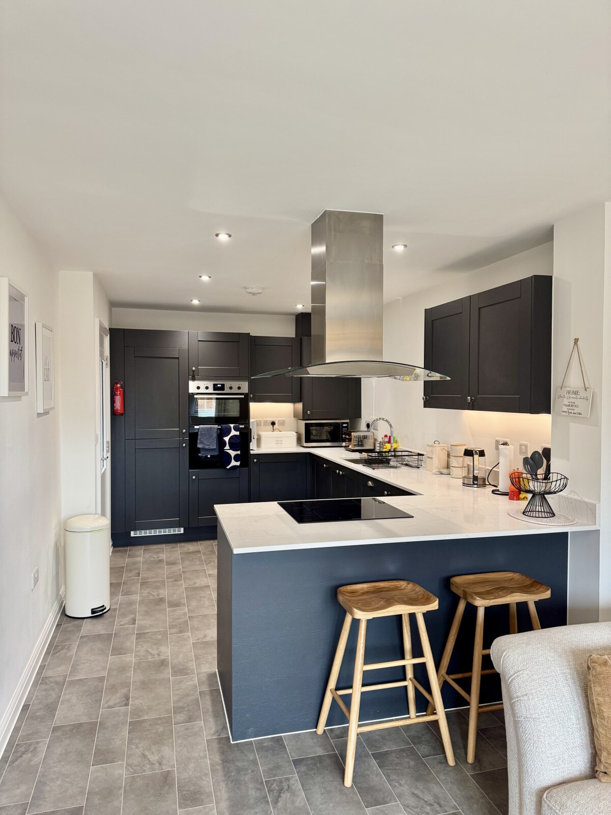 A modern kitchen features dark cabinetry and a sleek white countertop. Bar stools are positioned at a small breakfast bar. Stainless steel appliances, including an oven and microwave, are integrated into the cabinetry. Soft lighting illuminates the space, enhancing its functionality.