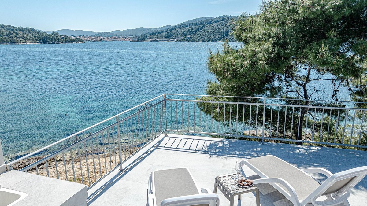 The spacious terrace overlooks the serene sea, providing unobstructed views of the water and distant hills. Two sun loungers are positioned comfortably on the deck, and a small table can be seen nearby, enhancing the outdoor relaxation space.