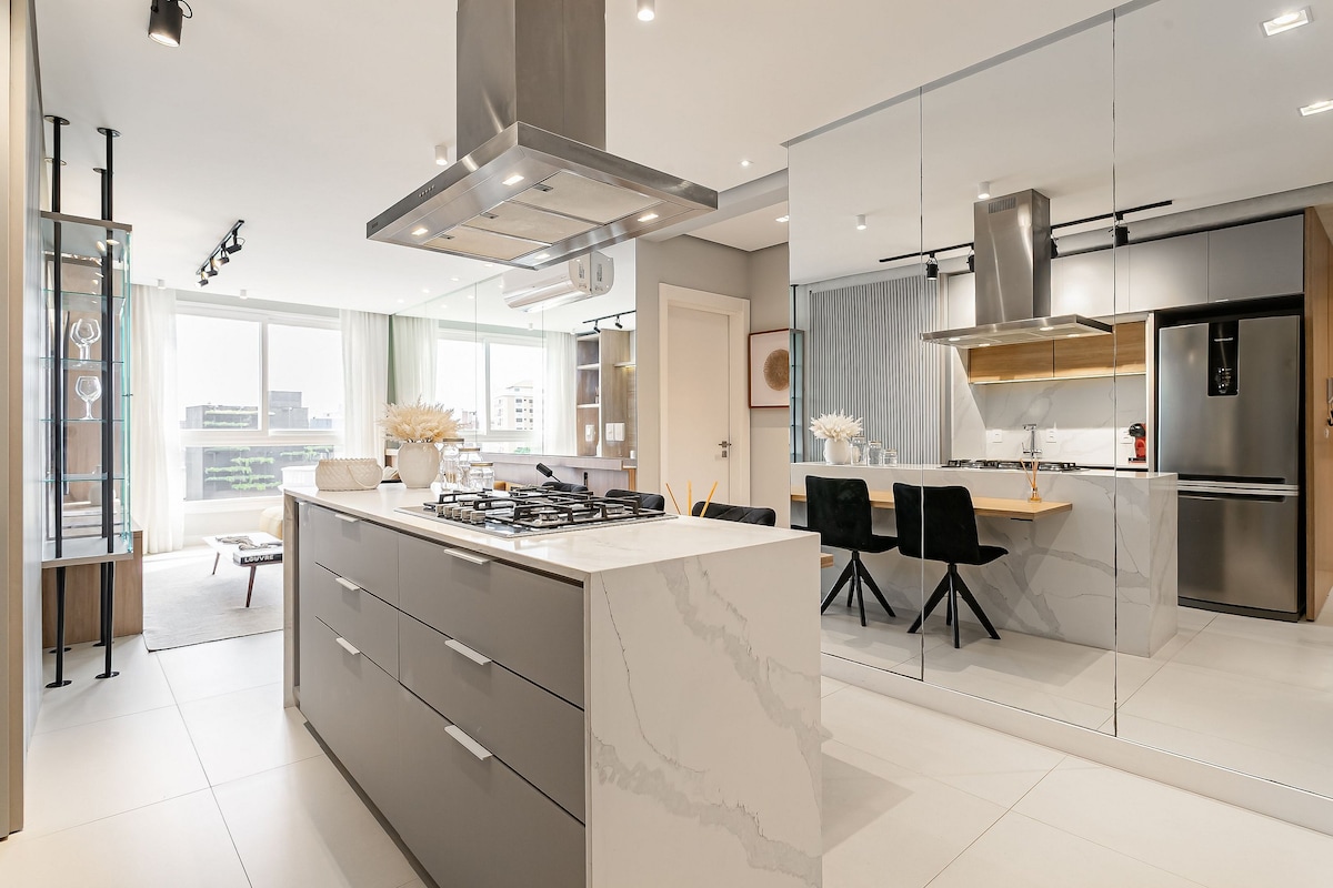 The open kitchen features a central island with a gas stove and modern cabinetry. A dining area with black upholstered chairs and a wooden table is visible in the background. Large windows allow natural light to illuminate the space, complemented by sleek, minimalist design elements.