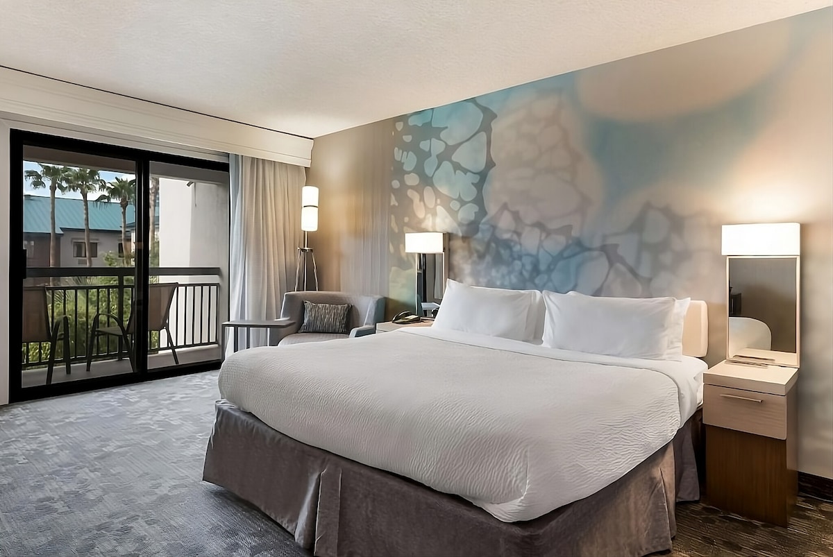 The room features a spacious king bed dressed in crisp white linens, complemented by a light grey bed skirt. A seating area is situated beside floor-to-ceiling glass doors that open to a private balcony. Soft lighting illuminates the contemporary decor, highlighted by an artistic wall mural.