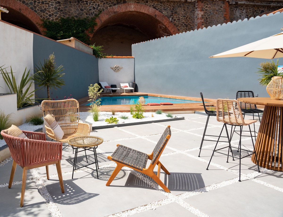 An outdoor lounge area features a mix of seating options, including woven chairs and a wooden table, arranged around a geometric stone patio. A small swimming pool is visible in the background, surrounded by greenery and modern design elements.