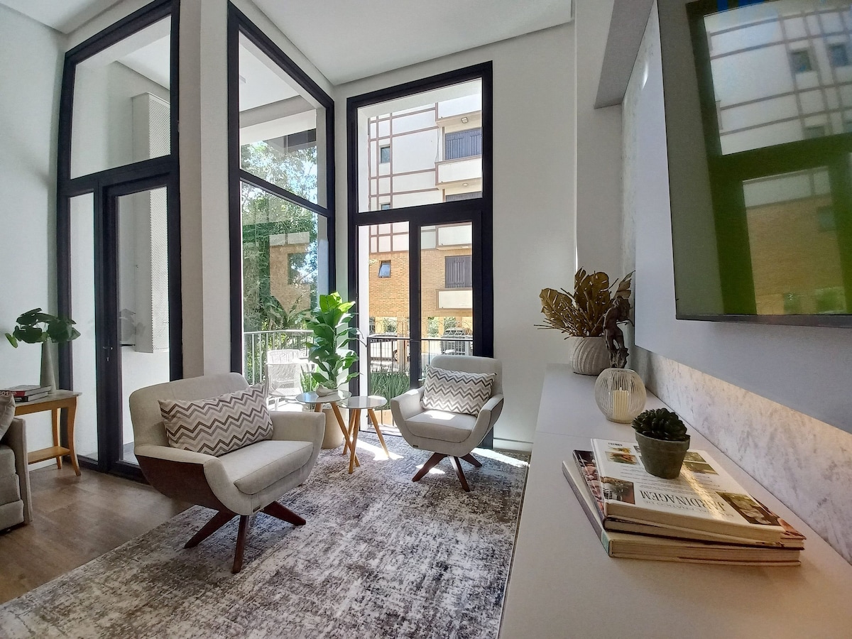 A modern living space is featured with large windows allowing natural light to fill the room. Two comfortable chairs with patterned cushions are positioned around a light rug. A console table displays decorative items, including a plant and books, creating a neat and inviting environment.