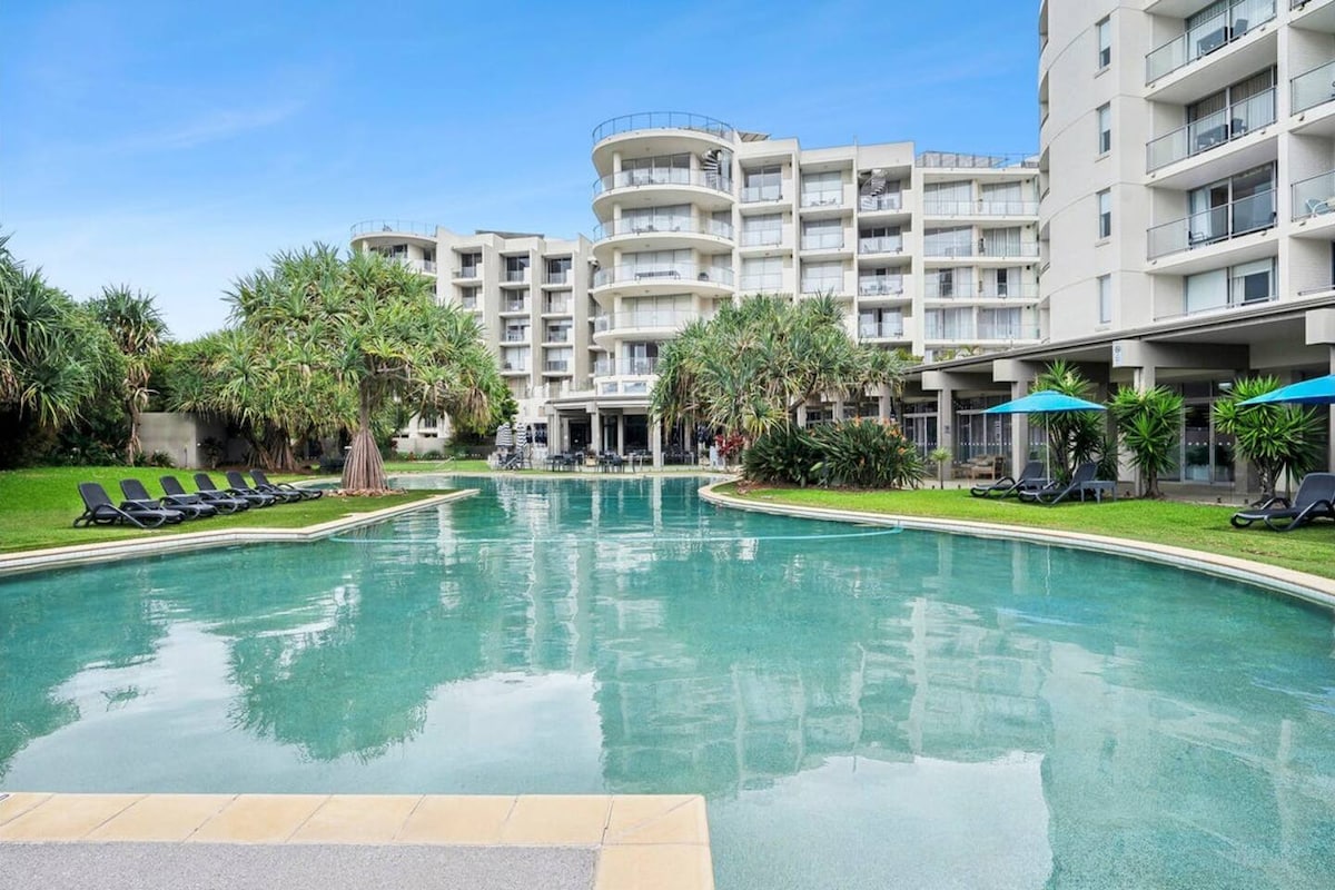 A lagoon-style pool is surrounded by lush greenery and tropical landscaping. The tranquil water reflects the building's modern architecture, while lounge chairs provide space for relaxation. The serene atmosphere invites guests to unwind in a peaceful setting.