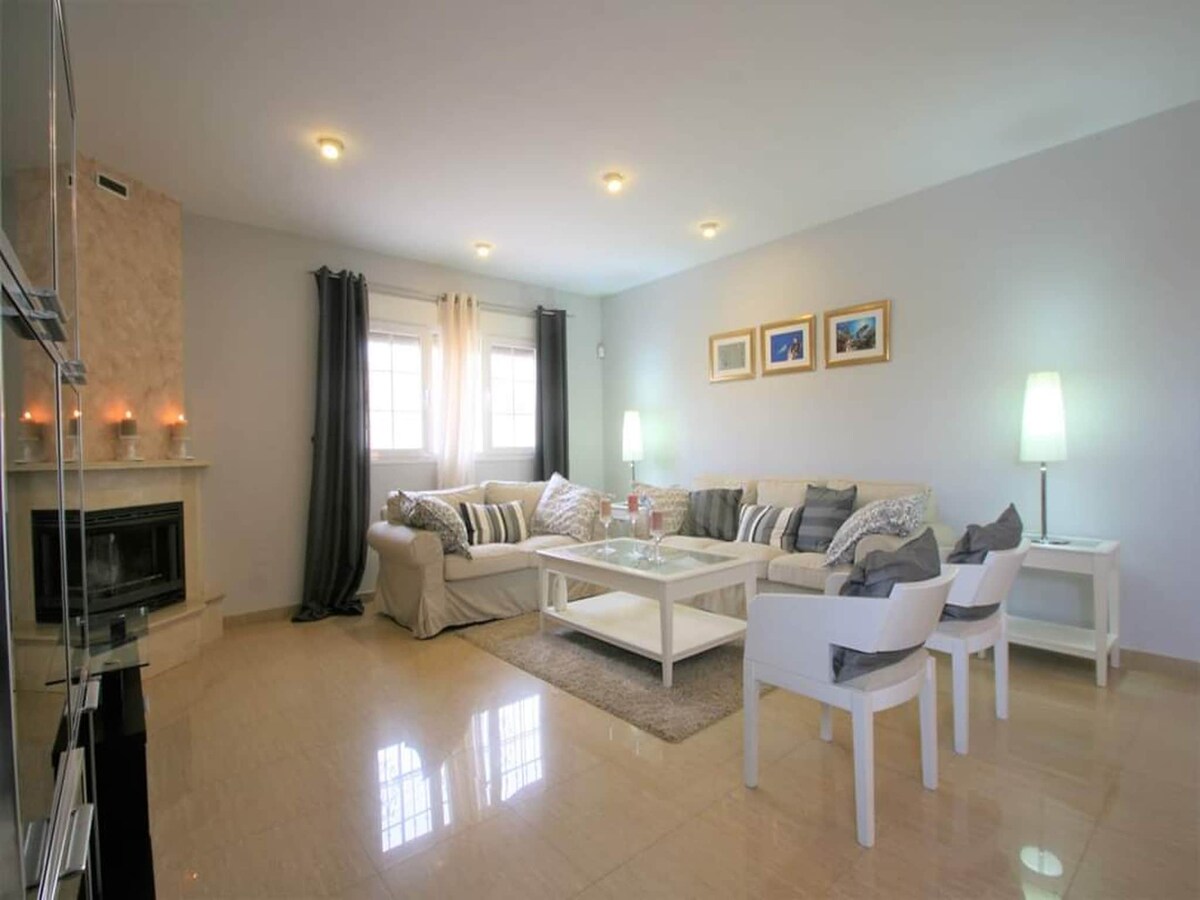 A bright living area is presented with a comfortable white sofa adorned with a variety of cushions. A coffee table sits in the center, surrounded by light seating. Natural light enters through multiple windows, while soft lighting fixtures add a calming ambiance.