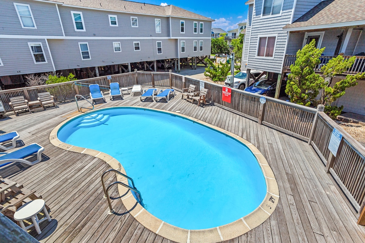 A community pool is depicted surrounded by a wooden deck featuring lounge chairs and tables. The pool's clear blue water reflects sunlight, while nearby buildings provide context to the pool's location within the community.