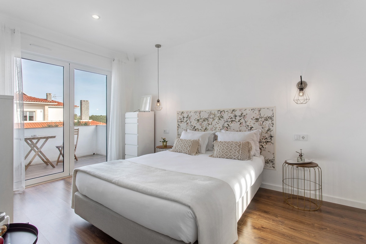 A bright bedroom features a spacious bed adorned with soft linens and decorative pillows. Natural light filters through glass doors that lead to a private balcony. A minimalist bedside table and a white dresser are visible, complementing the light wood flooring.