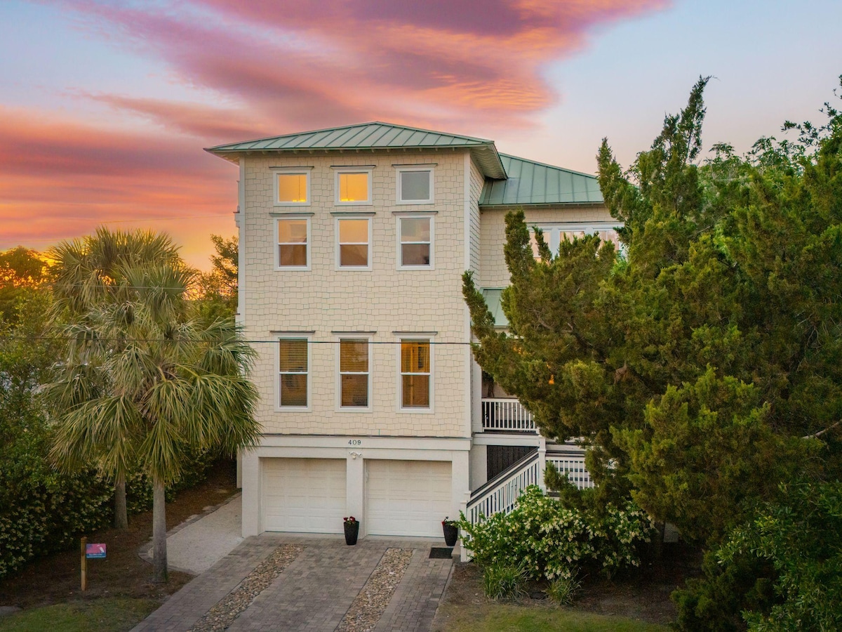 The exterior of a three-story beach home is presented, featuring a combination of light-colored siding and a green metal roof. Large windows are visible on the upper levels, reflecting the warm hues of sunset. Lush greenery surrounds the property, along with a driveway leading to the garage.