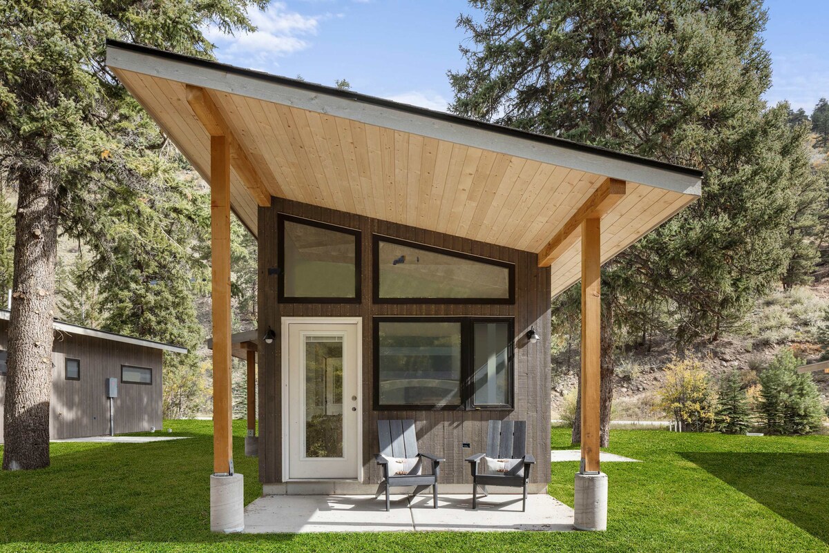 The cabin exterior is characterized by a modern architectural design, featuring an angled roof and large windows that invite natural light. Two chairs are positioned on a concrete patio, surrounded by vibrant green grass and trees, creating a peaceful outdoor setting.