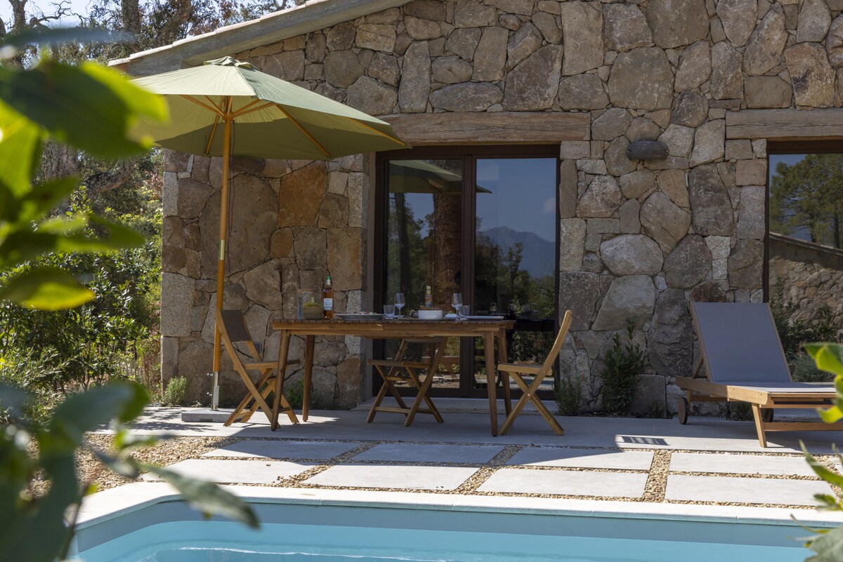 A serene terrace features a wooden dining table set with glasses and a bottle, shaded by a large green umbrella. A comfortable lounge chair is positioned nearby, while a private pool reflects the surroundings. The rustic stone exterior of the villa provides a natural backdrop.