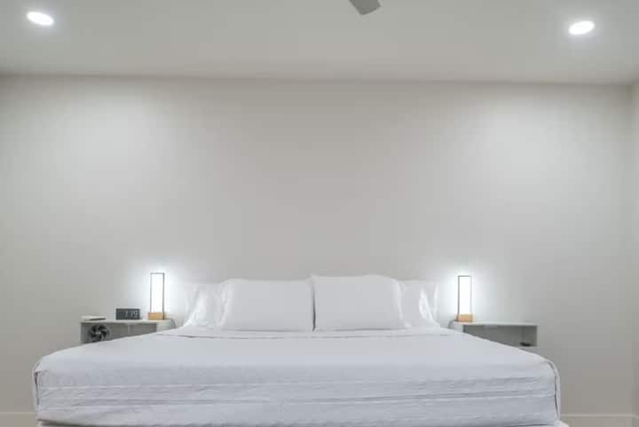 Minimalistická spálňa s pokojnou bielou estetikou s priestrannou manželskou posteľou, mäkkým osvetlením moderných nočných lámp a atmosférou bez neporiadku ideálnej na pokojné noci.