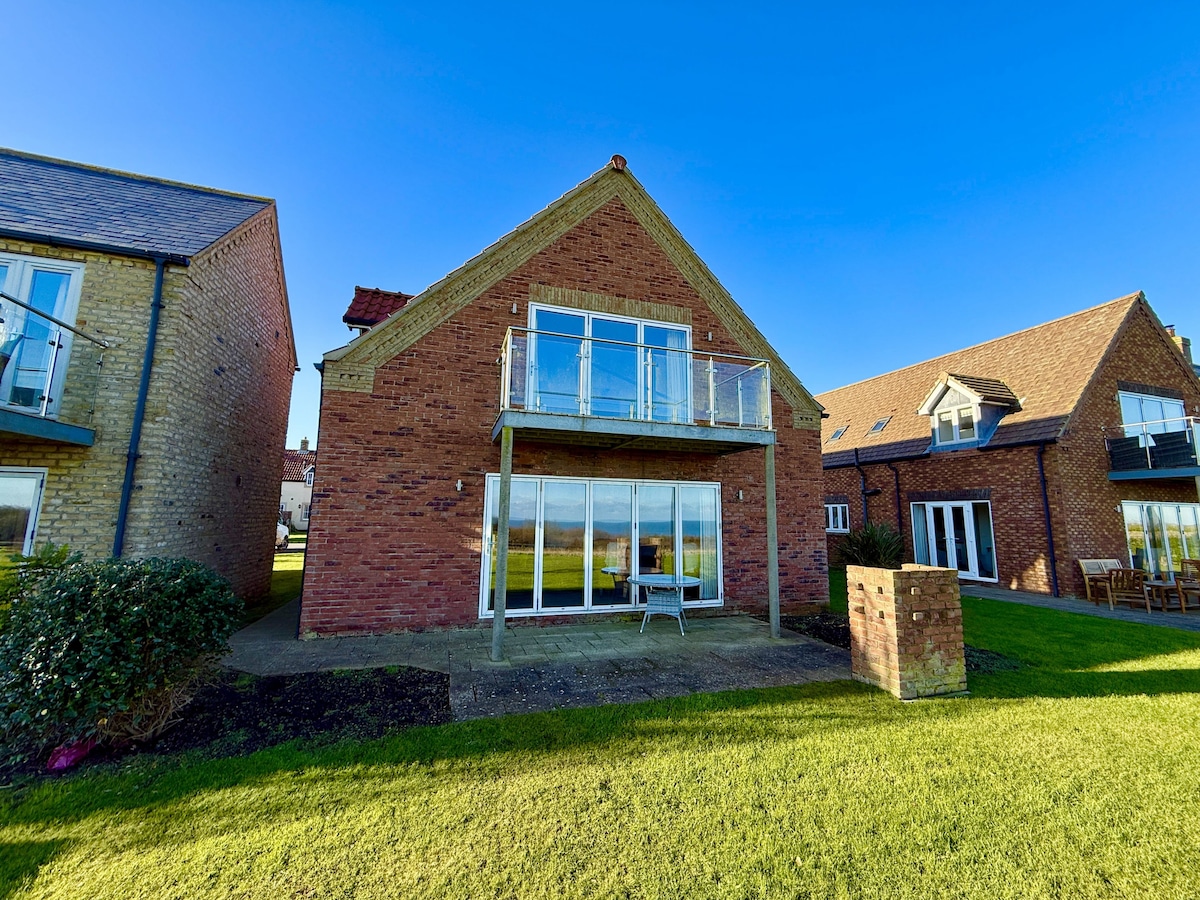 The exterior of the cottage is displayed, featuring a two-story brick façade with large glass doors that provide extensive natural light. A balcony extends from the upper level, offering views of the surroundings. Well-maintained grass and landscaping are present in the foreground.