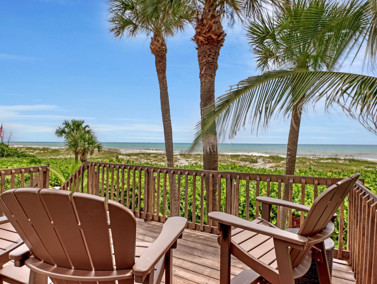 A shaded raised deck offers two adirondack chairs facing the ocean. Swaying palm trees frame the views of the sandy beach and blue water, allowing for a serene outdoor escape. The clear sky enhances the tranquil coastal atmosphere.