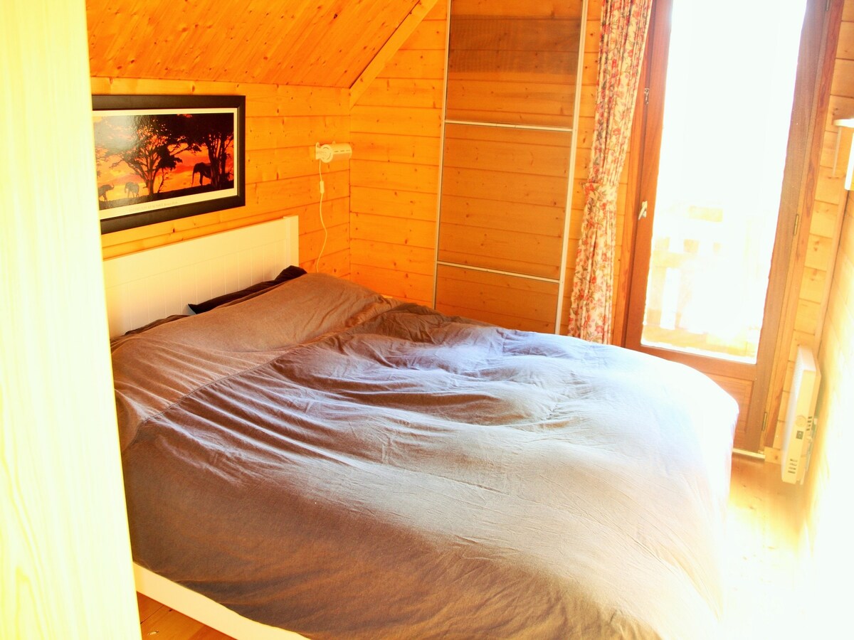 A spacious sleeping area features a large bed draped in soft, dark bedding. Natural light flows in from a window adorned with floral curtains, illuminating wooden walls. A piece of artwork is displayed above the bed, adding a touch of character to the room.