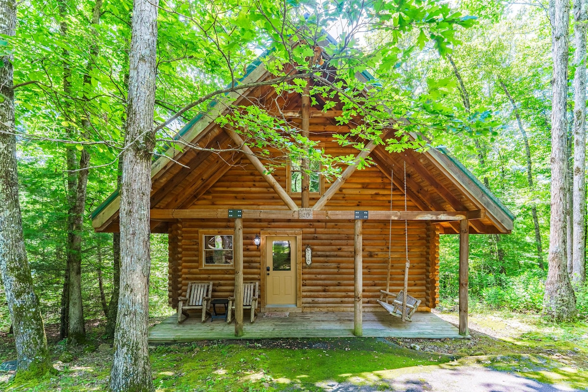The exterior of a log cabin is framed by lush green trees, offering a natural setting. A swing hangs from the porch, while two wooden chairs are positioned on the deck. The entrance features a simple door, inviting guests into the retreat.