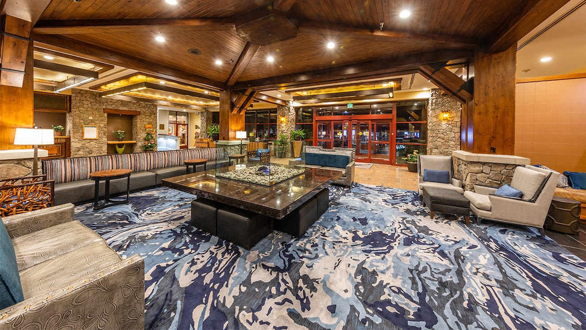 A spacious lobby is presented with wooden beams and stone accents. Comfortable seating is arranged around a low table set with decorative items. A vibrant area rug features a dynamic blue pattern, contrasting with the warm wood tones and providing a welcoming ambiance.