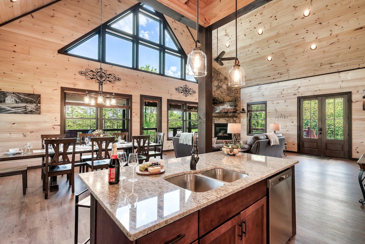 An inviting open-concept space features a modern kitchen with a large island and stainless steel sink. Natural light pours in through the tall windows, illuminating the wood-paneled walls and stylish dining area. A cozy living area is visible, showcasing a stone fireplace and comfortable seating.