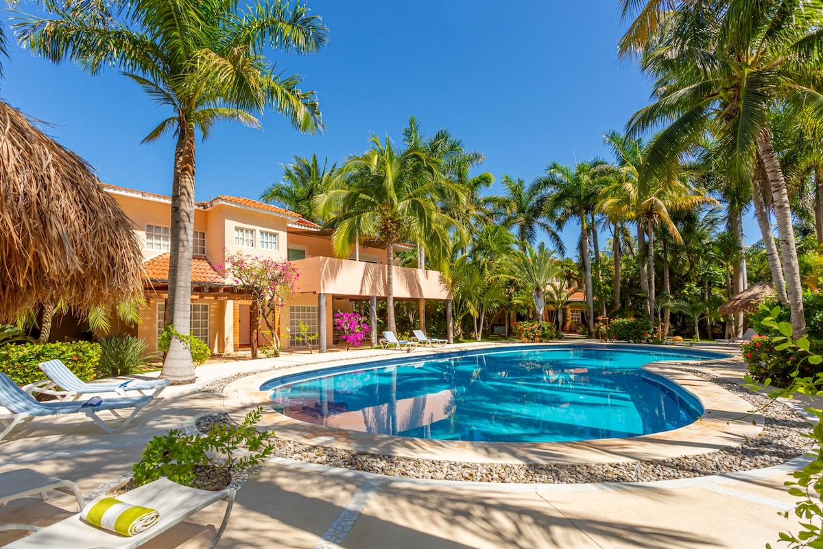 The outdoor area features a clear blue pool surrounded by lush palm trees and colorful blooming flowers. Lounge chairs are placed along the poolside, offering a space for relaxation under the sun. A thatched-roof gazebo provides shade nearby.