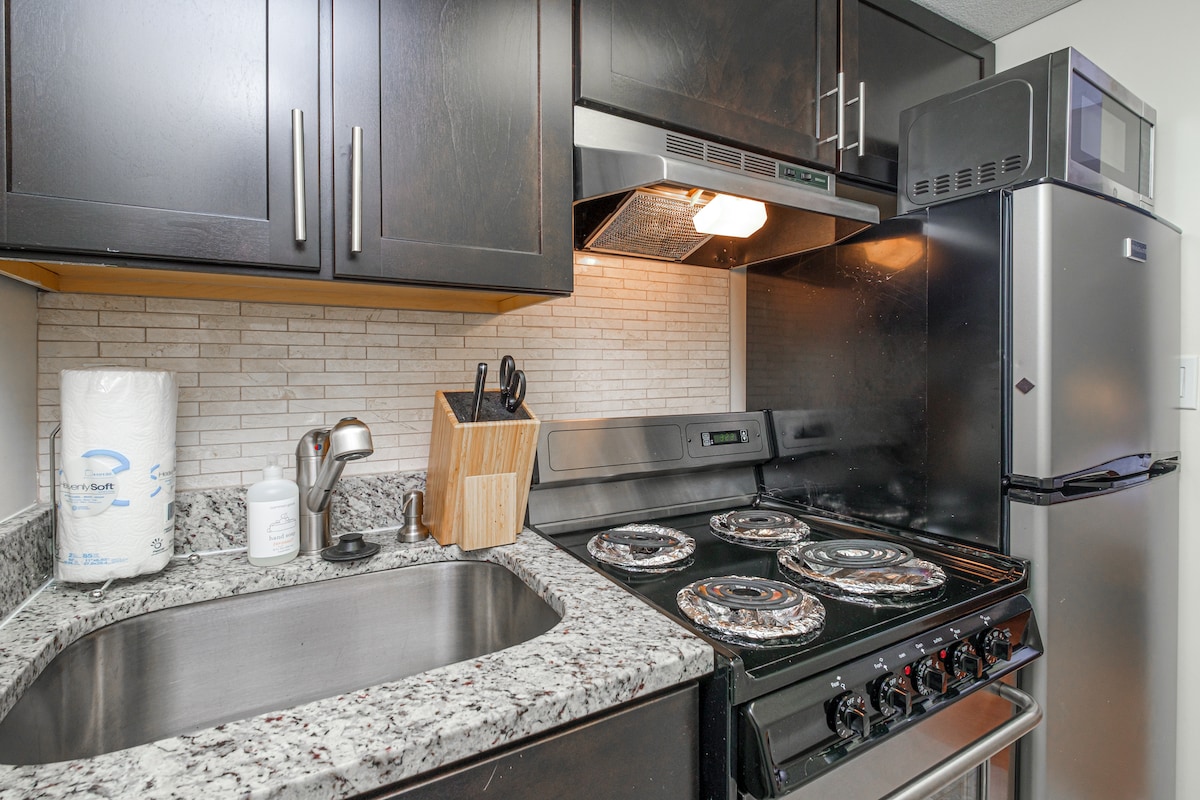 A fully equipped kitchen is displayed, featuring dark cabinets and a granite countertop. A stainless steel stove with four burners and a sleek vent hood are visible. Nearby, a microwave rests above a stainless steel refrigerator, alongside kitchen utensils organized in a wooden holder.