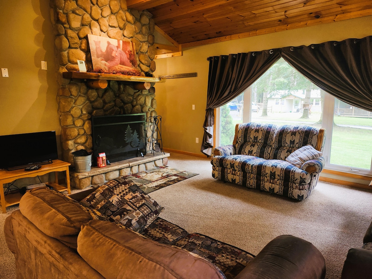 A spacious living room features a large stone wood-burning fireplace as the focal point. Two comfortable sofas are arranged for relaxation, while a television sits on a wooden stand. Large windows with drapes allow natural light, offering views of the surrounding area.