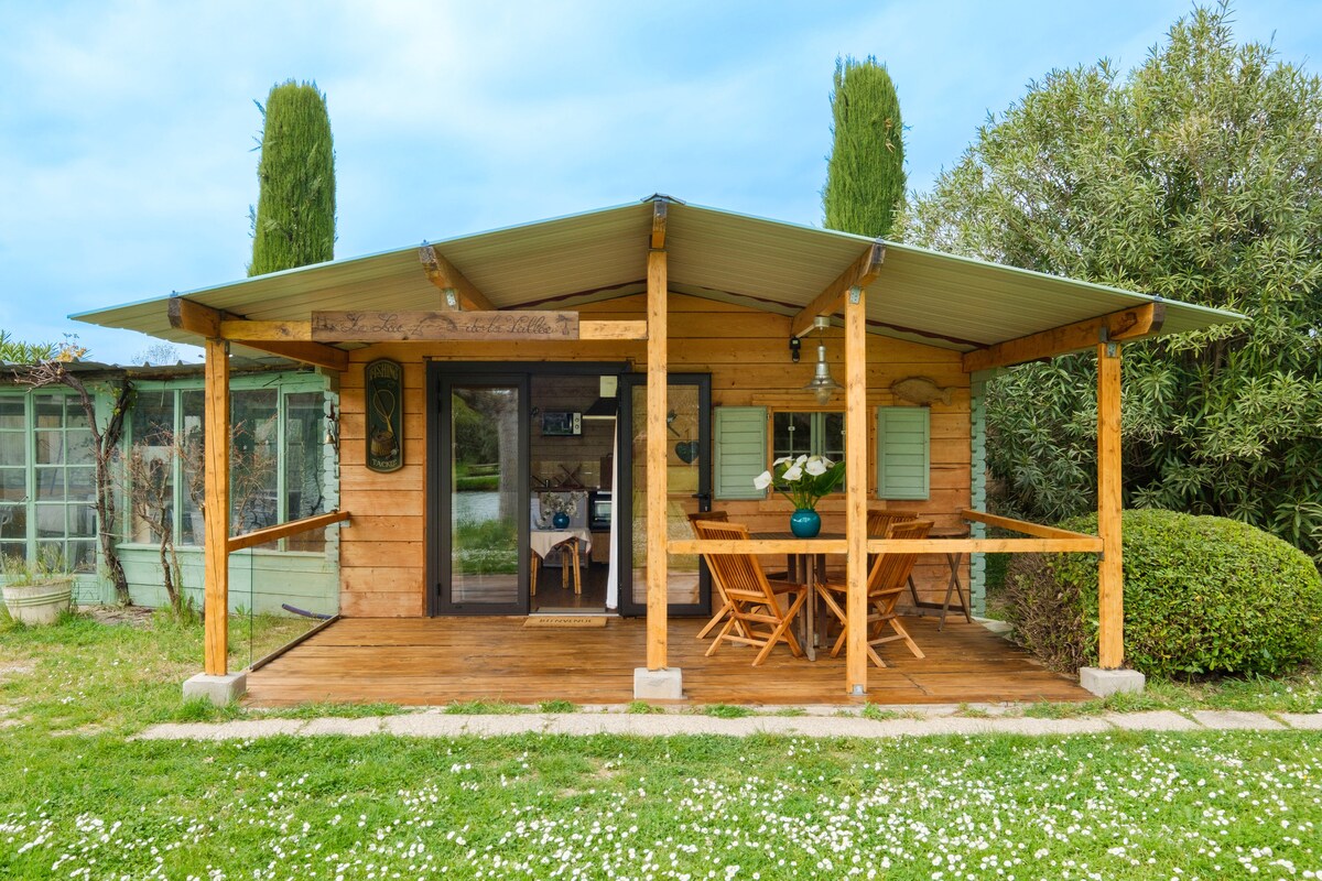 The front porch of the chalet is shown with a wooden deck and a large outdoor dining table surrounded by chairs. A refreshing view of the landscaped garden is visible, along with a glimpse of the pond in the background. The structure features a covered roof and wooden accents.