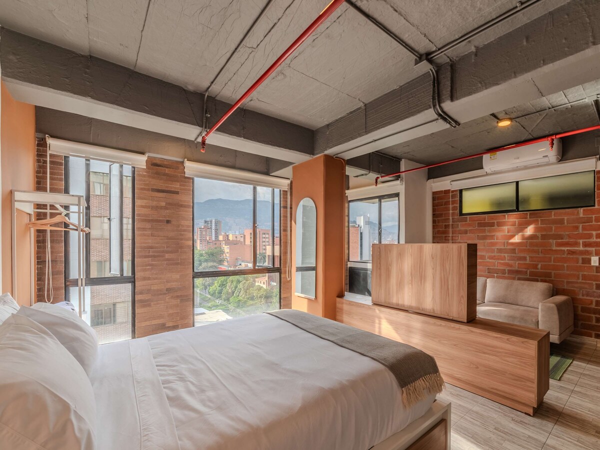 The interior boasts a modern bedroom with a spacious bed dressed in crisp linens. Large windows allow natural light to fill the space, showcasing views of the surrounding area. A cozy seating area is positioned nearby, with a blending of textures and colors.