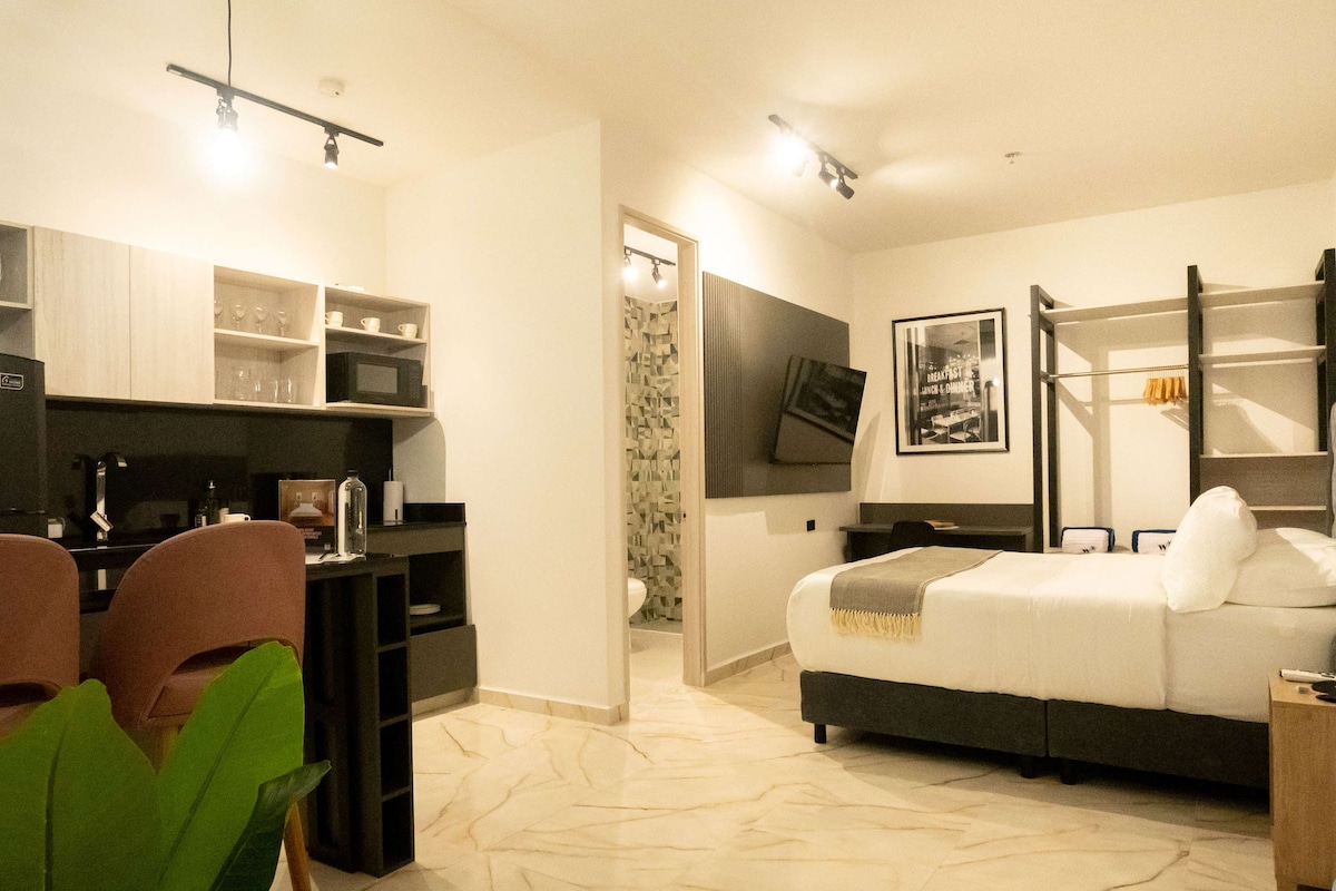 An inviting loft space features a queen bed and a stylish work area with two chairs. A fully equipped kitchenette is visible along with a modern bathroom entrance. Light-colored tile flooring and wall art enhance the ambiance, while flat-screen TV is mounted for convenience.