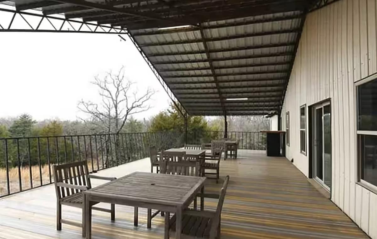 A spacious back deck features multiple wooden tables and chairs, providing ample seating for outdoor gatherings. The area is covered by a large overhang, offering protection from the elements, while scenic views of surrounding trees and open space are visible in the distance.