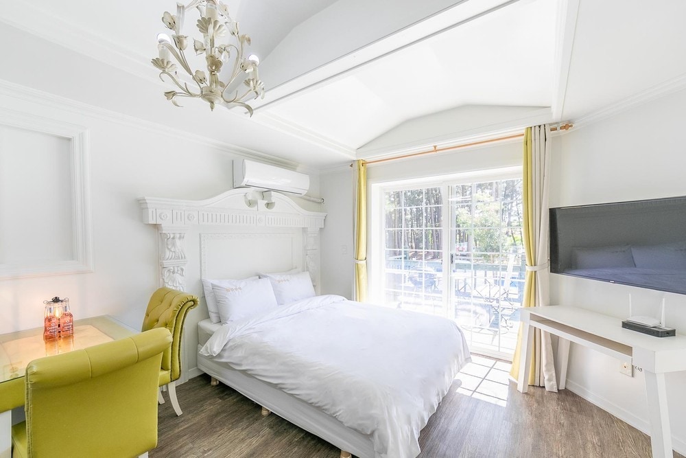 A bright bedroom showcases a spacious bed with crisp white linens, accompanied by a decorative chandelier overhead. A small table with two vibrant green chairs is positioned near a large window, inviting natural light into the room. A flat-screen TV is mounted on the opposite wall.