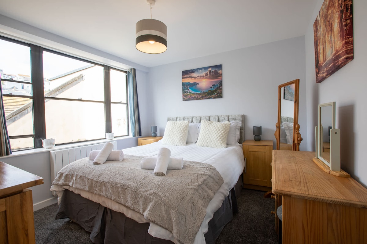 A well-lit bedroom features a spacious bed adorned with a light-toned blanket and decorative pillows. A large window provides natural light, framed by neutral curtains. Wooden furnishings, including a mirror and bedside tables, complement the decor, while a landscape art piece adds an inviting touch.