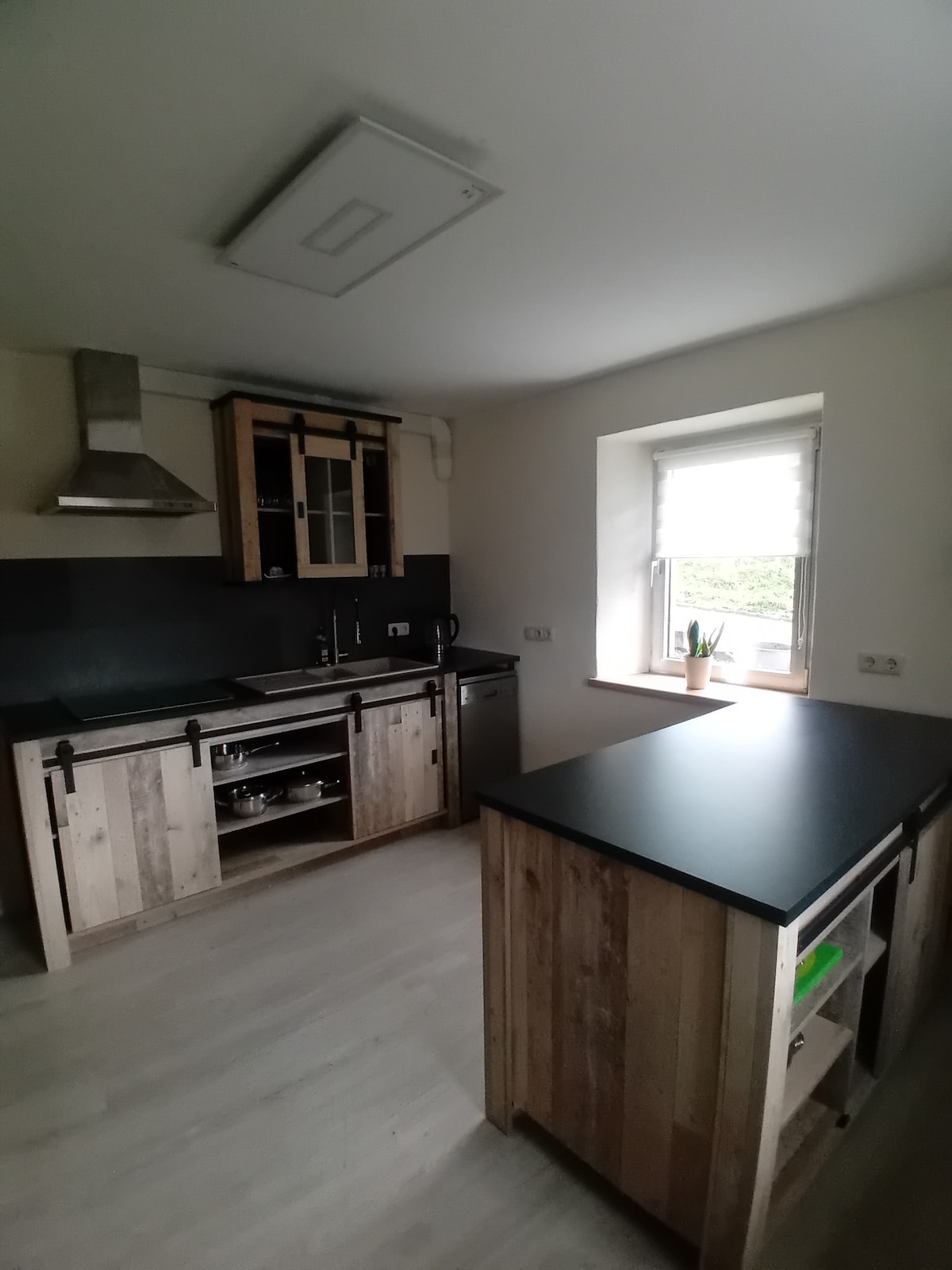 The kitchen features modern wooden cabinetry with a mix of open shelves and closed storage. A stainless-steel range hood is present above the cooking area. A window provides natural light, illuminating the workspace. Countertops are dark, and an island adds extra prep space.