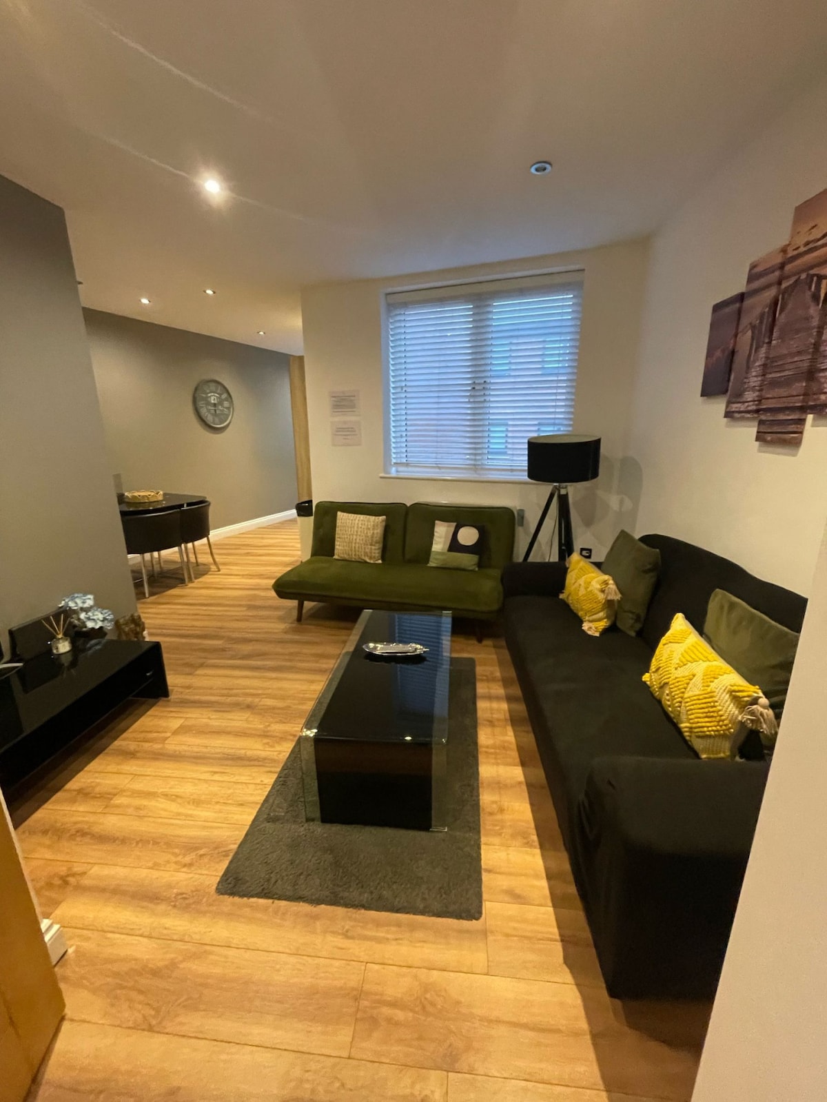 A cozy living area features a dark sofa adorned with yellow accent pillows, paired with a glass coffee table on a textured rug. A wall-mounted lamp and a decorative clock enhance the space, complemented by warm wood flooring and natural light filtering through the window.