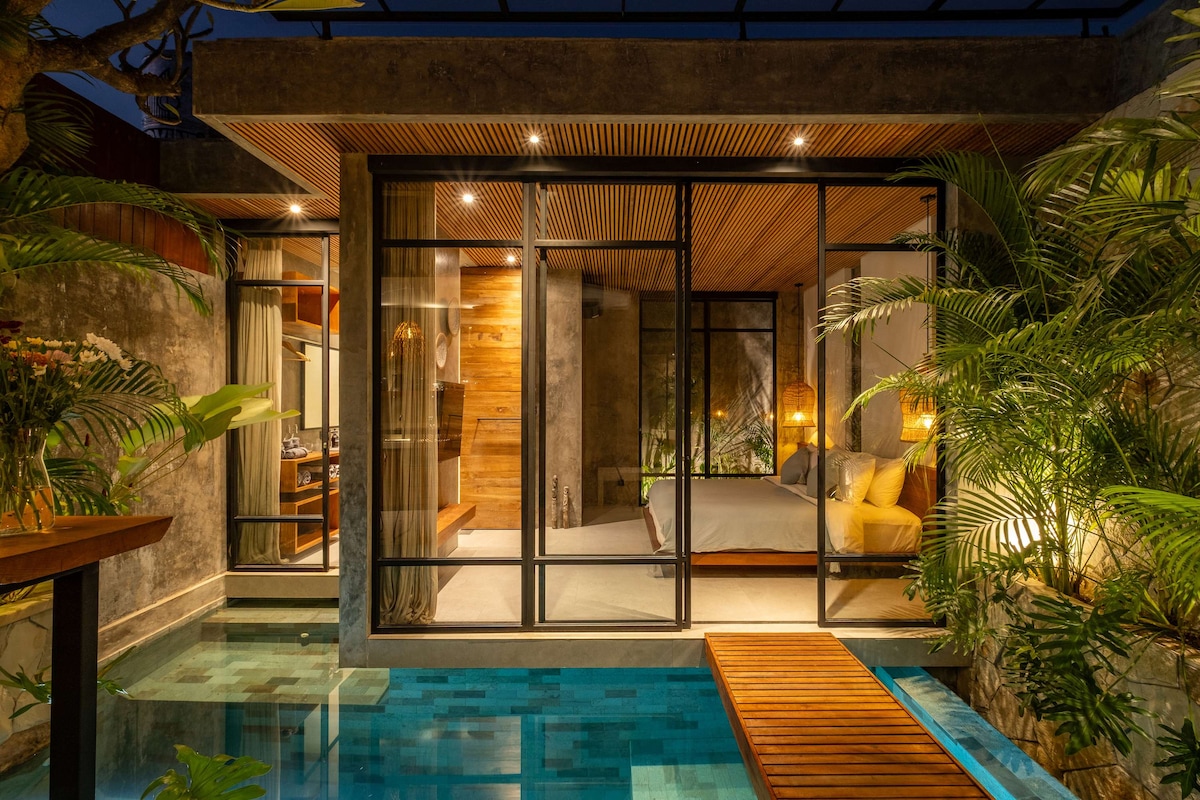 The villa is illuminated under the evening sky, showcasing expansive glass walls that connect the indoor living space to the private pool. Lush tropical plants surround the area, enhancing the serene atmosphere. A wooden pathway leads into the tranquil turquoise water of the private pool.