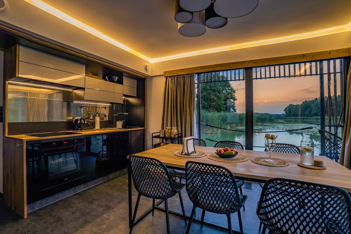 A modern kitchen and dining area are presented with sleek cabinetry and contemporary appliances. A wooden dining table is set with simple tableware, complemented by black mesh chairs. Large glass doors offer views of the serene natural surroundings and let in soft evening light.