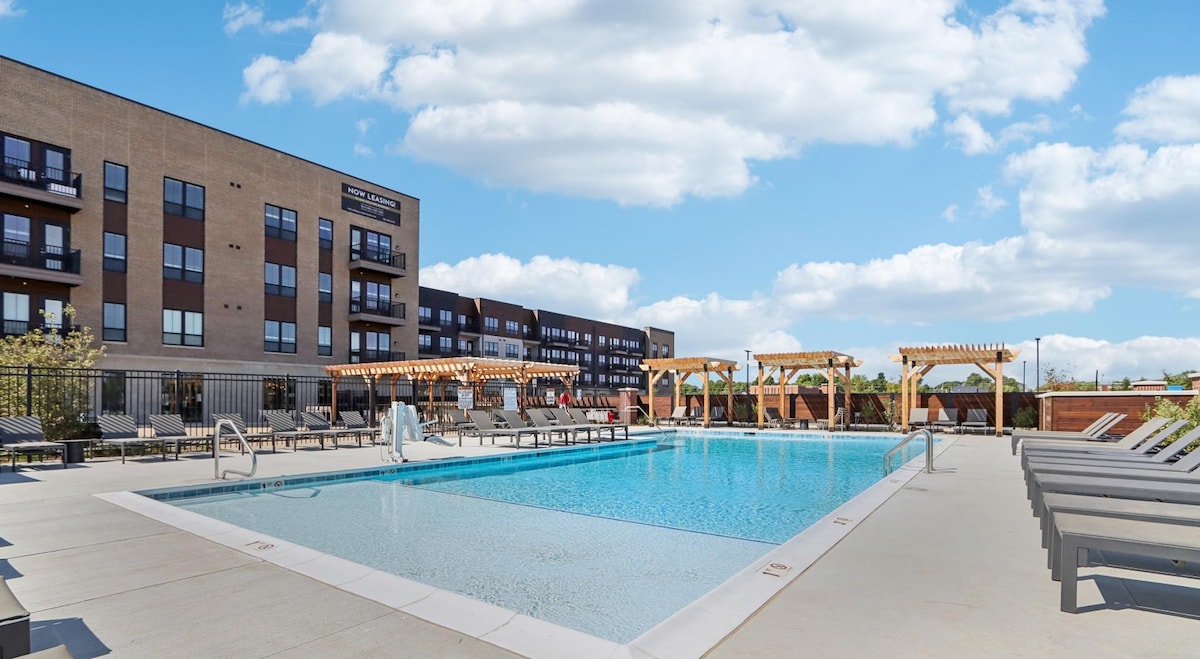 An outdoor swimming pool is surrounded by lounge chairs under a clear blue sky. Wooden pergolas provide shaded seating areas alongside the pool, enhancing the modern aesthetic of the community.
