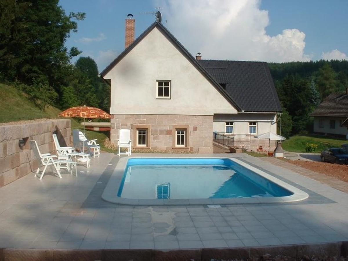 A large swimming pool is set in a stone-paved area adjacent to a modern holiday home. A few white lounge chairs are arranged nearby. The house features a pitched roof and multiple windows, surrounded by trees and gentle hills in the background.