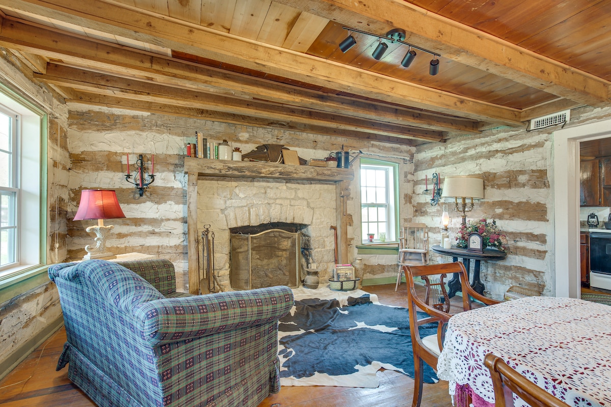 The living area features rustic log walls and wooden beams overhead. A stone fireplace acts as the focal point, flanked by windows that allow natural light to enter. A comfortable couch with a plaid pattern is paired with a wooden table and chairs, enhancing the cozy ambiance.