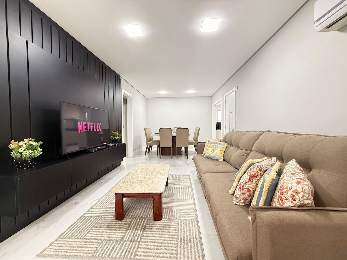 A spacious living room features a large, comfortable sofa adorned with decorative cushions. A coffee table sits in front of the sofa, and a wall-mounted television displays the Netflix logo. Behind, a dining area with a sizeable table for eight is visible.