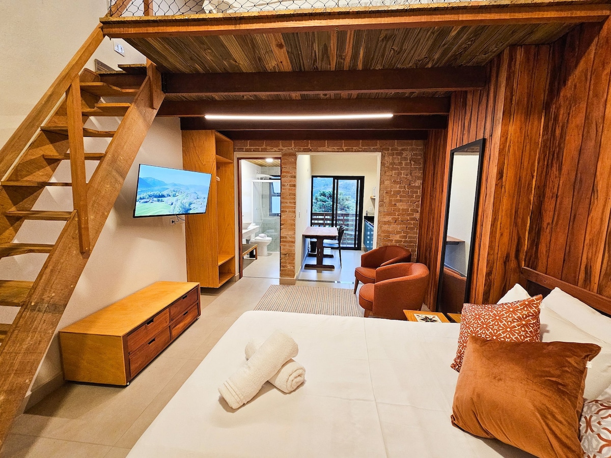 A modern interior is displayed with a spacious bed at the forefront, accompanied by neatly arranged towels. A wooden staircase leads to an upper level. Two armchairs are positioned adjacent to a low wooden cabinet, while a flat-screen TV is mounted on the wall, providing entertainment. Windows allow natural light to enhance the space.