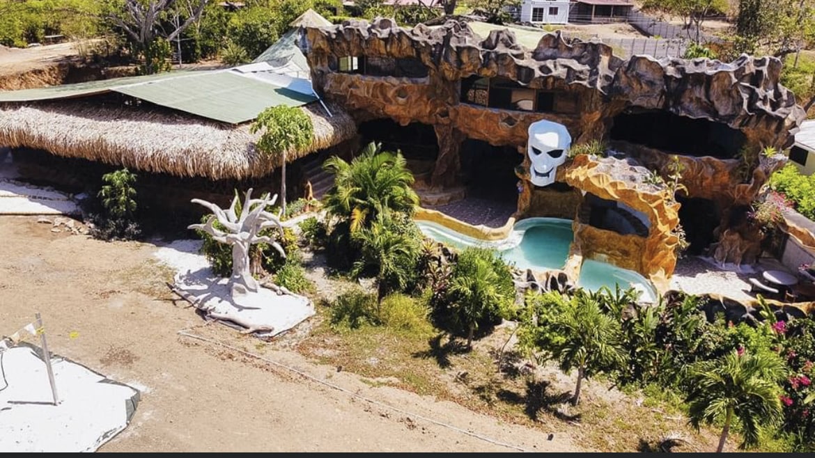 An aerial view of a distinctive structure characterized by stone textures and organic shapes, featuring a large skull motif. Surrounding the building, lush greenery and tropical plants create a vibrant landscape, while a small pool area invites relaxation under the sun. A thatched roof adds to the unique charm.