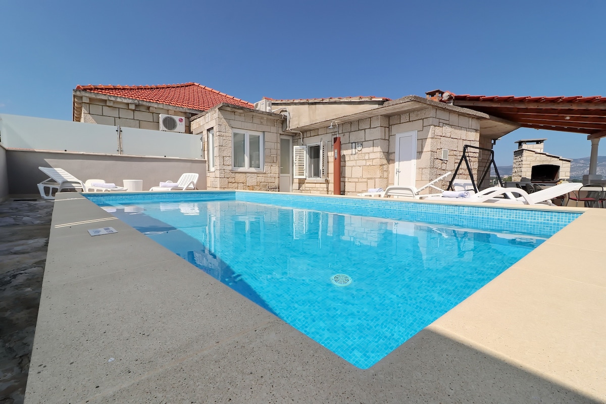 A private pool is surrounded by a spacious terrace furnished with sun beds and a swing chair. The villa's stone exterior and red-tiled roof are visible in the background, under a clear blue sky.
