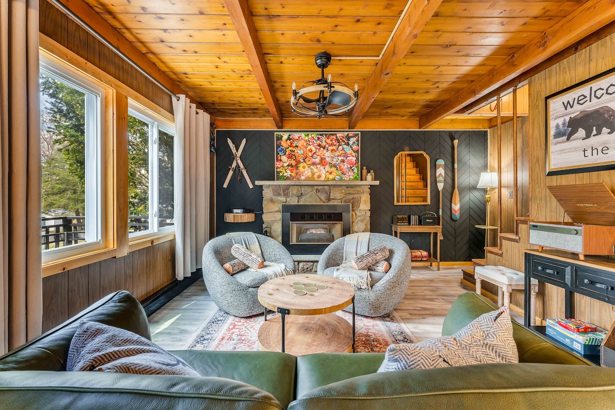 A cozy living area features a stone fireplace framed by two stylish armchairs and a circular wooden coffee table. Large windows allow natural light to fill the space, showcasing wooden beams on the ceiling and rustic decor elements, creating a welcoming atmosphere.