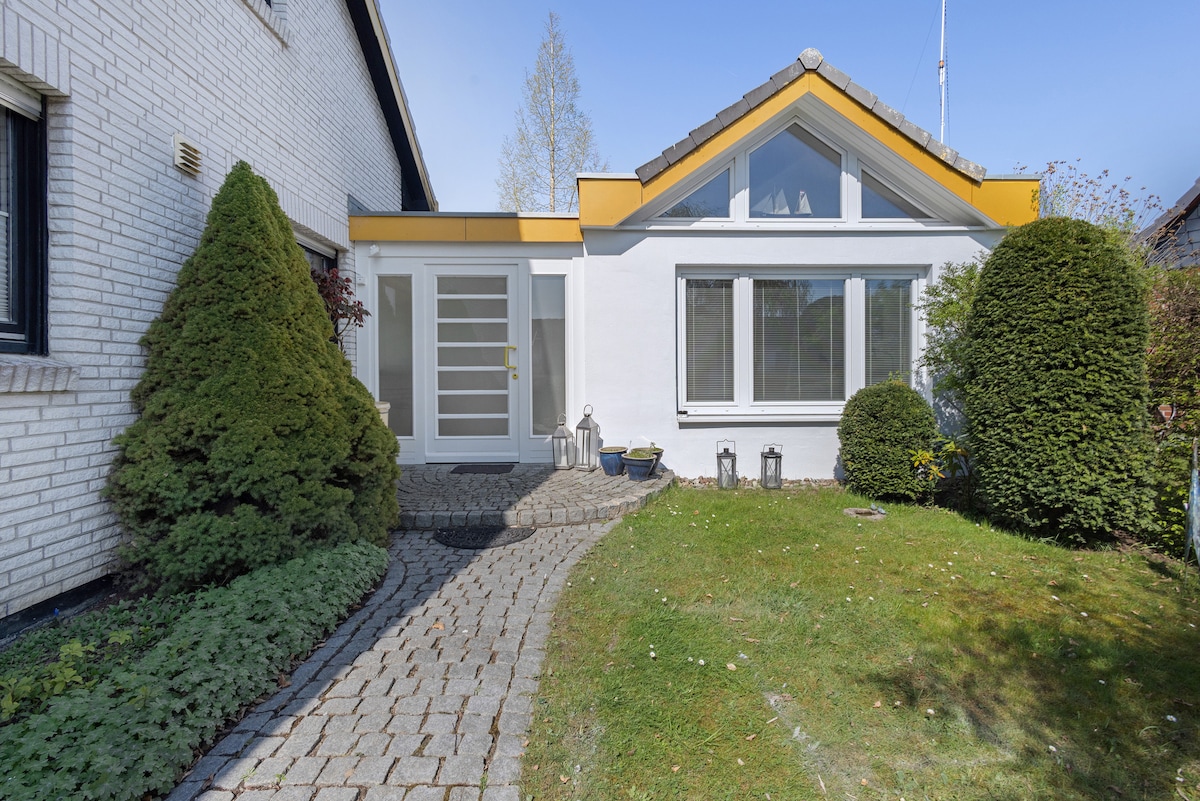 The exterior of the bungalow is shown, featuring a welcoming entrance with a modern door and large windows. Lush greenery, including well-trimmed bushes and a small garden, enhances the ambiance. A stone pathway leads to the entrance, bordered by a manicured lawn.