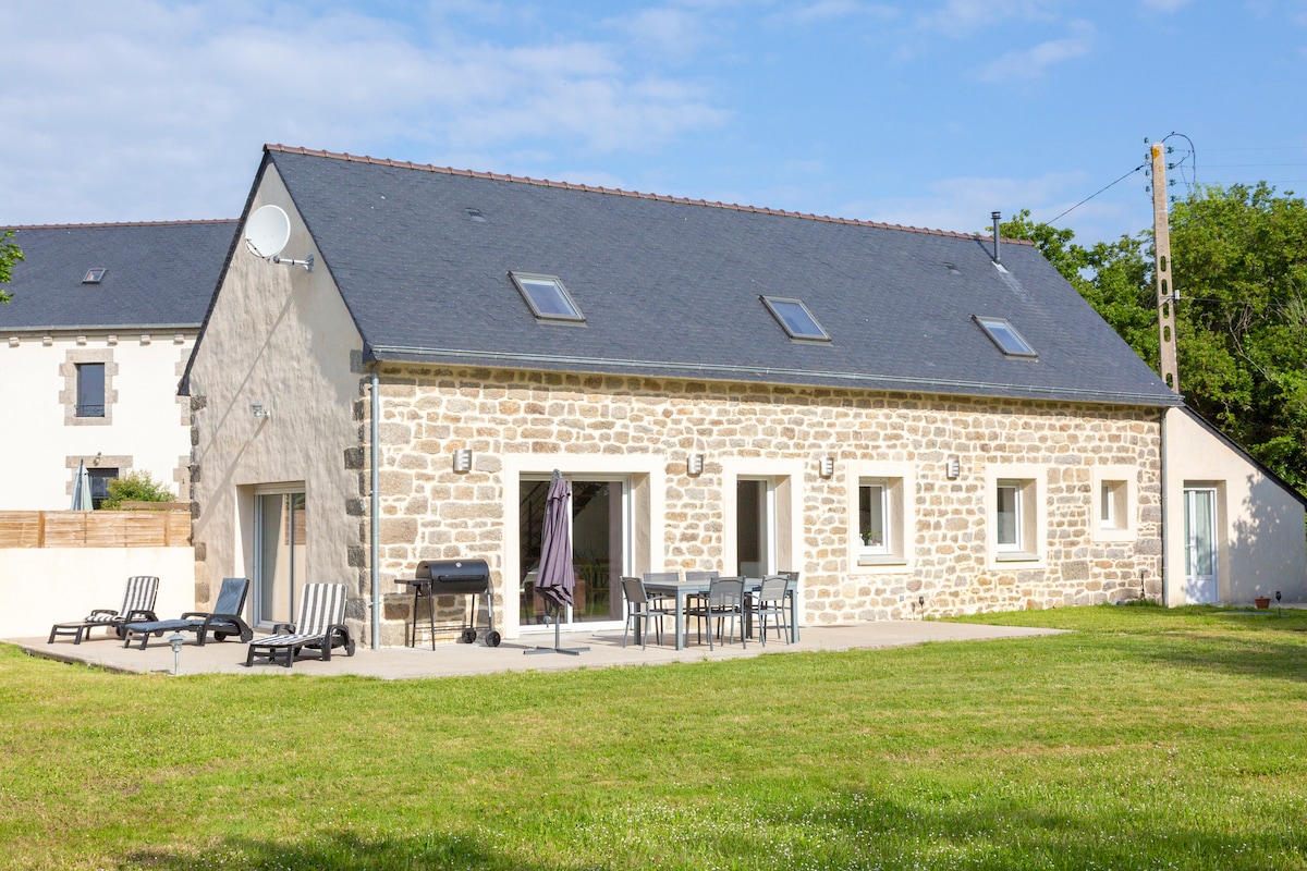 The exterior of a renovated farmhouse is displayed, featuring a light-colored stone façade and a slate roof. A spacious terrace with outdoor seating, including chairs and a dining table, is present. Lush green grass surrounds the area, and multiple windows allow for natural light.