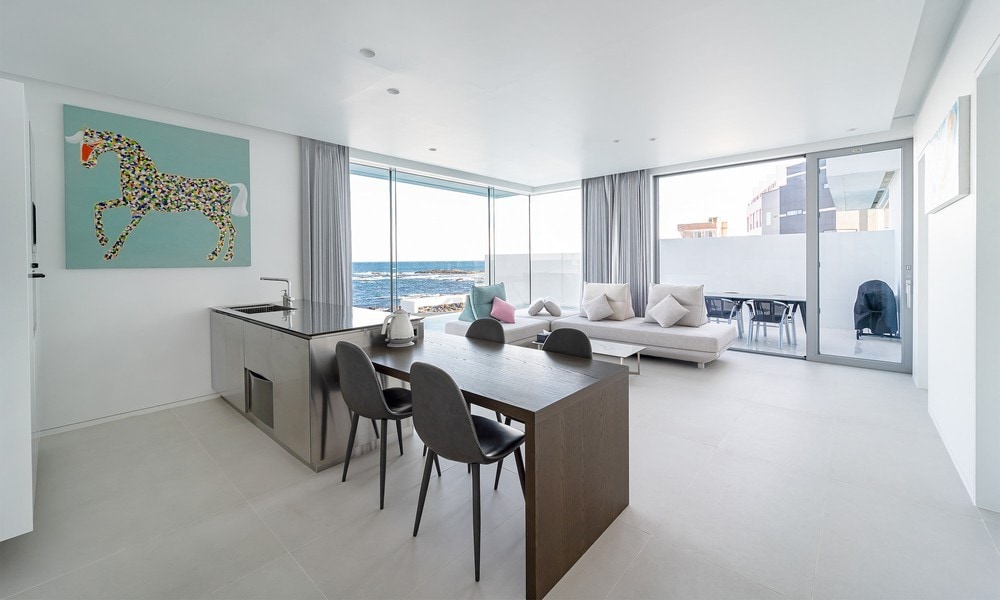 An open living area features a modern kitchen with a large dining table surrounded by four sleek chairs. Floor-to-ceiling windows offer ocean views, while light curtains frame the space. A lounge area with neutral couches is visible in the background.