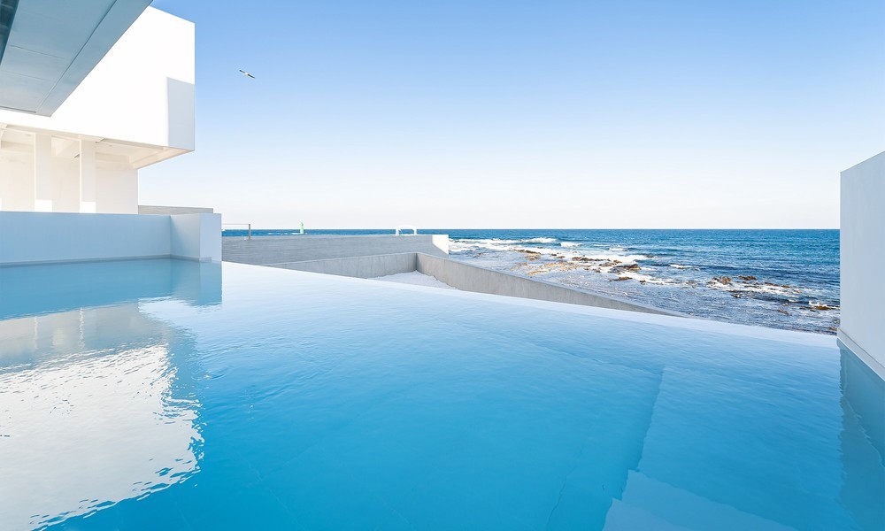 An infinity pool extends towards the ocean, creating a seamless view of the sky and water. The pool's surface reflects the clear blue sky, while the coastal rocks can be seen in the distance, enhancing the tranquil atmosphere of the outdoor space.