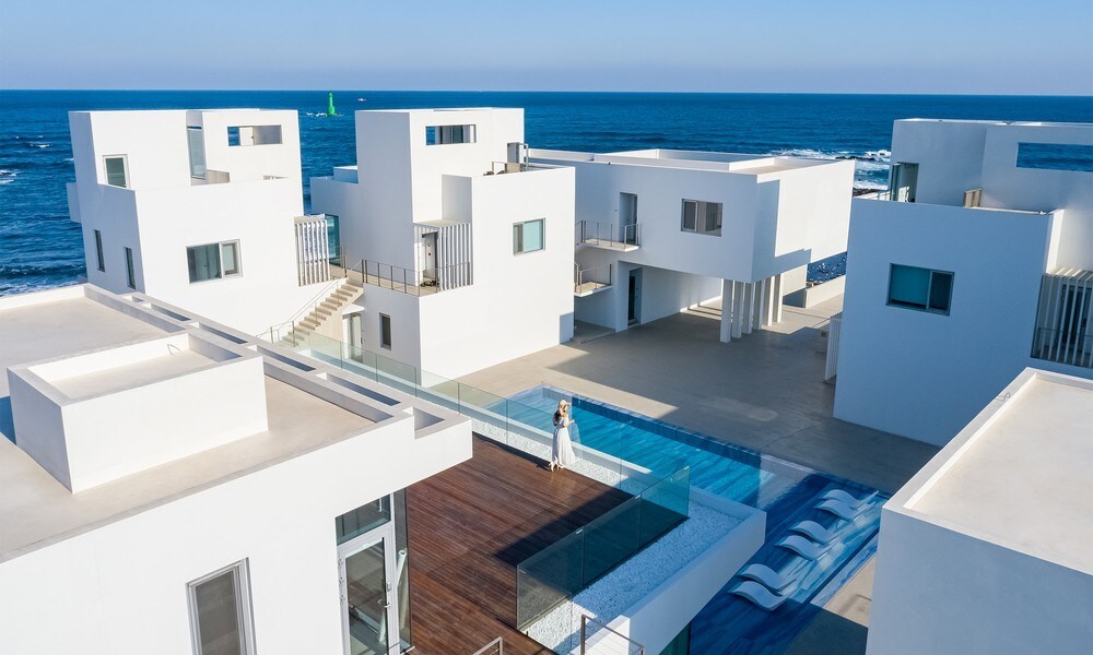 A modern villa complex is visible, featuring clean lines and minimalist white architecture. A spacious outdoor area with a pool runs through the center, bordered by lounge chairs. The expansive ocean can be seen in the background, enhancing the scenic coastal view.