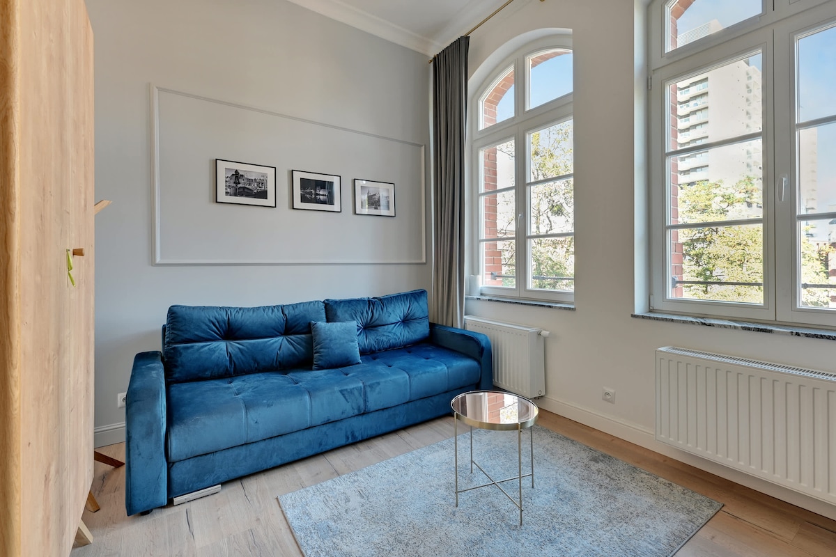 Un estudio luminoso y acogedor con un sofá azul, ventanas arqueadas y obras de arte enmarcadas que agregan un toque decorativo.